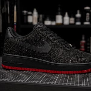 May include: Black and red Air Force 1 sneaker. The shoe features a black upper with a glittery texture, a black swoosh, and a black sole. The sole has a red trim. The sneaker is displayed on a black grid surface.