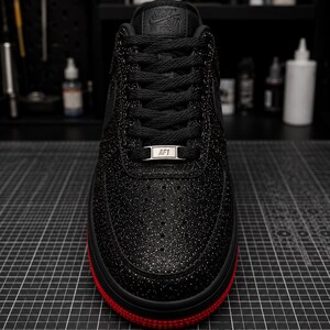 May include: Black Nike Air Force 1 sneaker with a glittery finish and red sole. The shoe features black laces, the Nike Air logo on the tongue, and an AF1 tag. The sneaker is on a black grid surface.