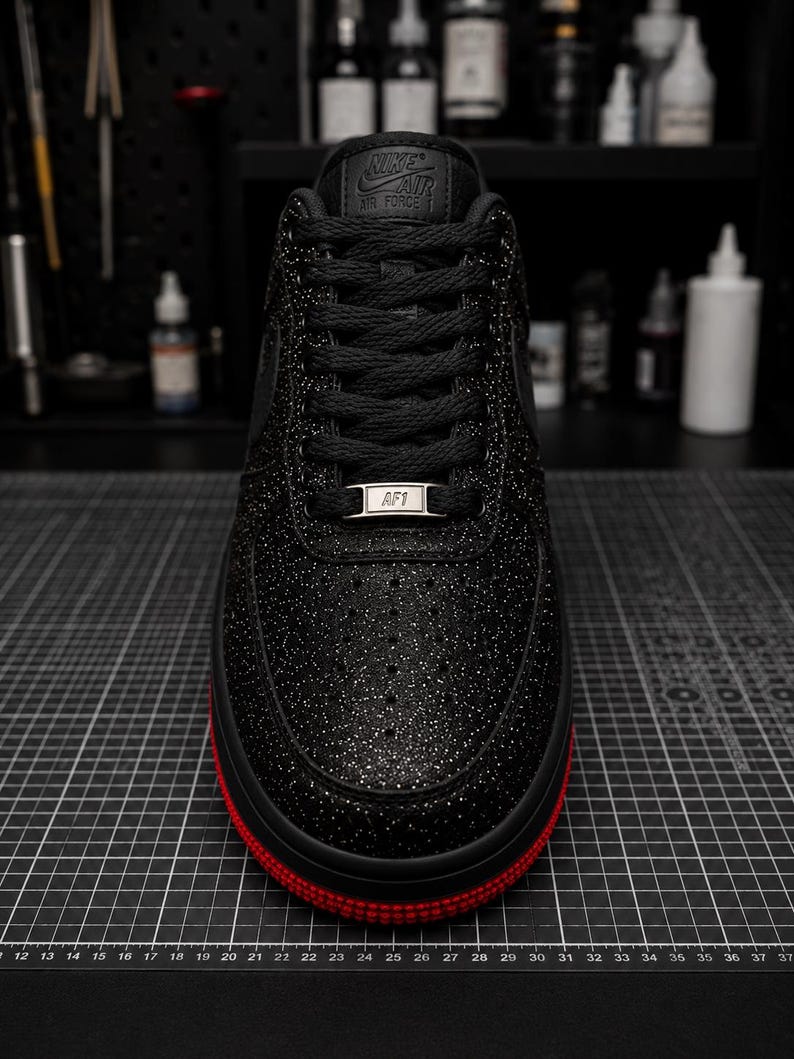 May include: Black Nike Air Force 1 sneaker with a glittery finish and red sole. The shoe features black laces, the Nike Air logo on the tongue, and an AF1 tag. The sneaker is on a black grid surface.