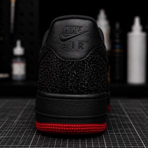 May include: Black Nike Air Force 1 sneaker with a red sole. The heel features the Nike logo and Air text. The shoe has a textured black upper with a speckled pattern.