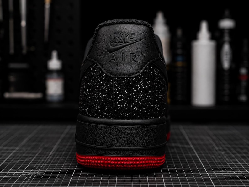 May include: Black Nike Air Force 1 sneaker with a red sole. The heel features the Nike logo and Air text. The shoe has a textured black upper with a speckled pattern.