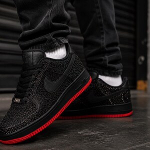 May include: Black sneakers with a textured upper and a red sole. The Nike swoosh is visible on the side. The shoes are worn with black jeans and white socks.