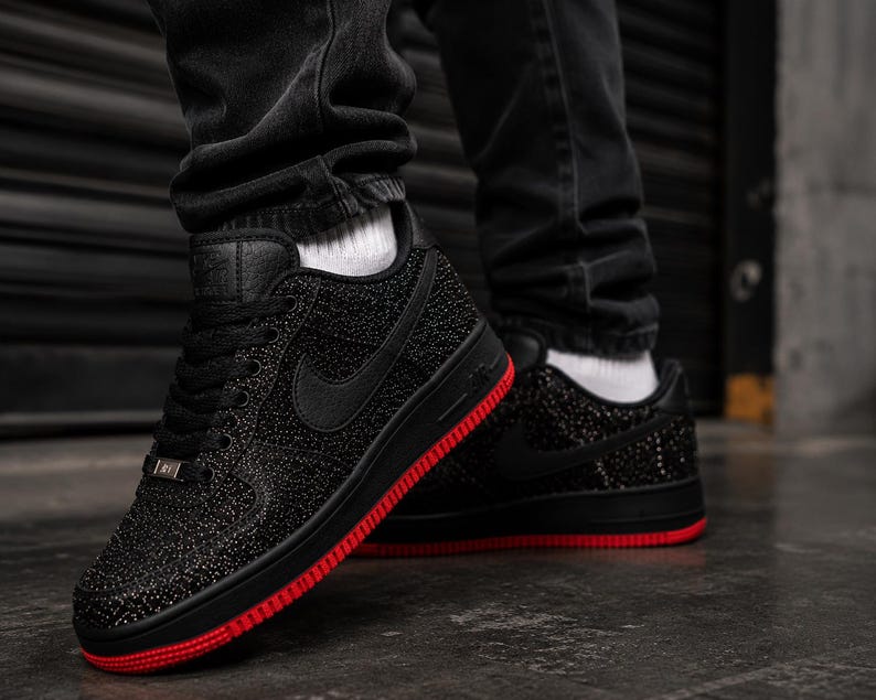 May include: Black sneakers with a textured upper and a red sole. The Nike swoosh is visible on the side. The shoes are worn with black jeans and white socks.