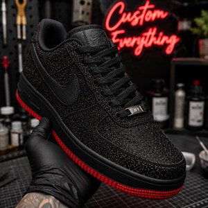 May include: Black Nike Air Force 1 sneakers with a red sole and a textured upper. The shoe features a black Nike swoosh and laces. The words "Custom Everything" are in red neon in the background.