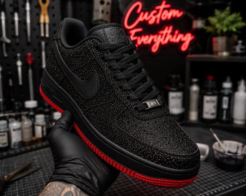 May include: Black Nike Air Force 1 sneakers with a red sole and a textured upper. The shoe features a black Nike swoosh and laces. The words "Custom Everything" are in red neon in the background.