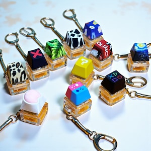 Könnte beinhalten: Eine Sammlung von Schlüsselanhängern mit bunten, gemusterten Tastenkappen. Designs umfassen einen Kuhdruck, ein Fragezeichen, eine Sternennacht und Spielkonsolen-Symbole. Jeder Schlüsselanhänger hat eine transparente, rechteckige Basis und eine silberne Kette.