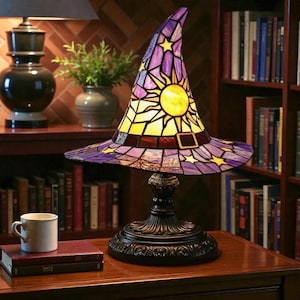 May include: A stained glass table lamp in the shape of a witch's hat. The hat features a yellow sun and stars against a purple background. The lamp has a black, ornate base and sits on a wooden surface.