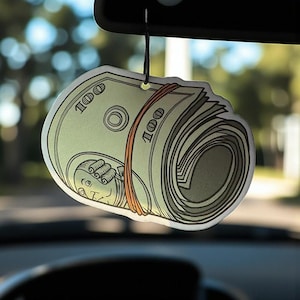 Op de afbeelding: Luchtverfrisser voor in de auto in de vorm van een opgerolde stapel van 100 dollar biljetten, vastgehouden door een bruine elastiek. Het hangt aan een zwart touwtje. De achtergrond toont een achteruitkijkspiegel en een vage buitenomgeving.
