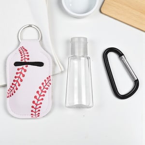 May include: A white baseball-themed hand sanitizer holder with red stitching detail and a silver keyring. A clear, empty bottle and a black carabiner clip are also present. The items are arranged on a white surface.