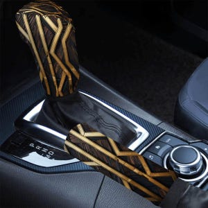 May include: A car gear shift knob cover and a parking brake handle cover featuring a dark brown wood grain pattern overlaid with a geometric design of intersecting gold lines. The covers are installed on a vehicle center console.