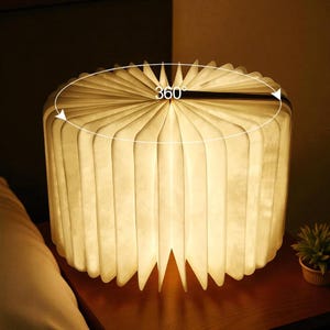 May include: A glowing, round lamp with a book-like design. The lamp is made of white, folded paper that emits a warm light. The text "360°" is visible on the top. The lamp sits on a wooden surface, next to a small potted plant.