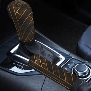 May include: A car gear shift knob cover and a handbrake lever cover featuring a dark brown wood grain pattern with intersecting gold geometric lines. Both covers are fitted over the interior controls of a vehicle with a black console.