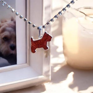 Peut inclure: Un collier argenté avec un pendentif en forme de chien. Le pendentif est de couleur brun-rouge avec un contour argenté. La chaîne du collier est également argentée et ornée de petits cristaux transparents. Une photo encadrée d'un chien est en arrière-plan.