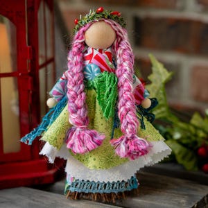 May include: A handmade doll with a wooden head, long pink braided hair, and a green and white patterned dress. The doll has a floral crown and is decorated with blue and green accents. The doll is approximately 7 inches tall.