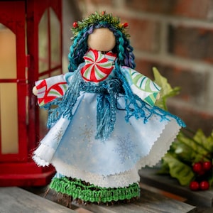 May include: A handmade Christmas doll with a fabric body, wearing a light blue dress with snowflake patterns and a white lace trim. The doll has blue yarn hair, a green wreath, and a peppermint candy design on the chest.