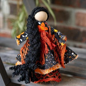 May include: A handmade doll with a wooden head, black yarn hair, and a black, orange, and white patterned dress. The doll has a long black braid and an orange crocheted shawl. The doll is standing on a wooden surface.