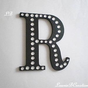 May include: Black wooden letter R with white rhinestones around the edges. The letter is a decorative piece for a home or nursery.
