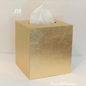 May include: A square, gold tissue box with a textured surface. White tissues are pulled from the top opening. The box has a metallic finish and is sitting on a white surface. The text "LB" is in the upper left corner.