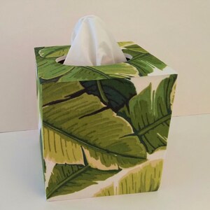 K&ouml;nnte beinhalten: Ein quadratischer Taschentuchbox-Bezug mit einem tropischen Bananenblatt-Druck in Gr&uuml;n- und Wei&szlig;t&ouml;nen.
