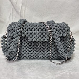 Könnte beinhalten: Graue, perlenbesetzte Handtasche mit silberner Kette. Die Tasche ist rechteckig mit abgerundeten Kanten und einem Klappenverschluss. Die Perlen sind klein und gleichmäßig verteilt, was der Tasche ein strukturiertes Aussehen verleiht. Der Hintergrund ist ein weicher, weißer Stoff.