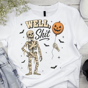 May include: A white t-shirt featuring a graphic of a mummy skeleton holding a jack-o'-lantern balloon. The text "WELL, Shit" is printed above the skeleton in a stylised font. Small black bats and star shapes surround the central illustration.
