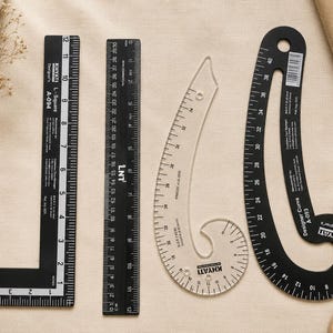 May include: A set of four sewing rulers including a black L-square ruler, a black straight ruler, a clear curved ruler, and a black curved ruler. Each tool features white printed measurement markings in inches and centimeters for pattern drafting.