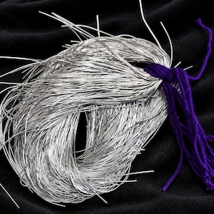 May include: A bundle of thin, flexible silver-coloured metallic wires gathered into a loose coil. The wires are secured at one end by a thick, vibrant purple yarn tie, with the individual wire ends extending outward in various directions.