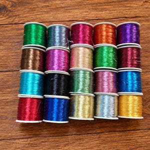 May include: A collection of twenty small spools of metallic embroidery thread arranged in a four-by-five grid. The spools feature a variety of vibrant colors including green, blue, red, purple, gold, silver, pink, and black with a shiny finish.