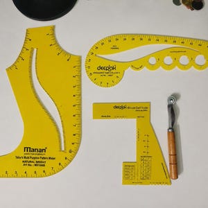 Puede incluir: Un conjunto de herramientas de costura de plástico amarillo que incluye un patrón multiusos Manan, una plantilla de diseño Deepak, una escala para pinzas de blusa Deepak y una rueda de trazado de metal con mango de madera, con marcas impresas.