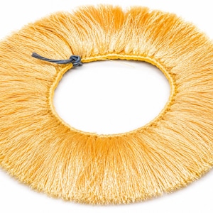 May include: A circular yellow fringe trim featuring a dense, silky texture with fine, straight threads radiating outward from a central stitched band. A small, dark grey cord is tied in a knot at the top edge of the circular trim.