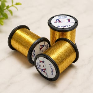 May include: Three spools of metallic gold embroidery thread with black plastic ends. Each spool features a white circular label printed with the text Special Quality A-1 MAMTA. The thread is wound tightly around the center of each spool.