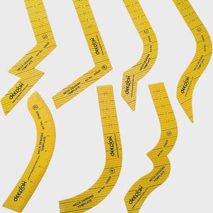 May include: A set of seven yellow plastic neck design templates labelled with the brand name Deepak, Art No. D5029, and various letter identifiers. Each template features printed measurement markings in inches and distinct curved or angular shapes.