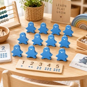 May include: A set of twelve blue wooden penguin-shaped figures arranged on a wooden table. The set includes a wooden number board with digits one through five, a wooden abacus, a wooden rainbow stacker, and educational cards with numbers and text.