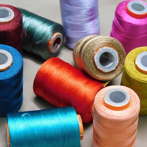 Puede incluir: Una colección de diez carretes cilíndricos de hilo de coser en varios colores vibrantes, incluyendo rojo oscuro, verde azulado, lavanda, magenta, azul, naranja, oro, amarillo, turquesa y melocotón, cada uno con un núcleo central de plástico blanco.