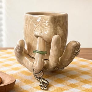 Puede incluir: Una taza de cerámica beige con forma de rostro, sostenida por una mano que sostiene un anillo verde y un collar. La taza descansa sobre una tela a cuadros amarillos, con galletas en un plato de madera. Un calendario muestra el 15 de septiembre.