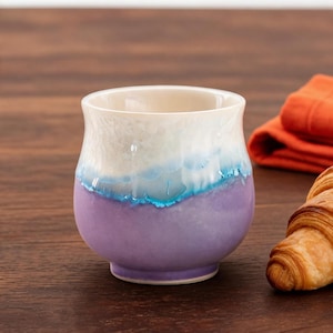 May include: A handmade ceramic cup with a white, blue, and purple glaze. A golden croissant and an orange napkin are next to the cup on a wooden surface. The cup is a unique piece of art.