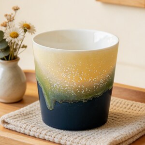 Puede incluir: Una taza de cerámica con un diseño degradado, que transiciona de blanco a amarillo, verde y azul marino. La taza está sobre un posavasos beige dentro de una bandeja de madera. Un pequeño jarrón con flores y la palabra "COZY" en un letrero de madera están en el fondo.