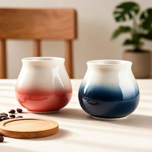 May include: Two ceramic coffee mugs with a gradient color design. One mug transitions from white to red, the other from white to blue. A wooden coaster and coffee beans are also visible.