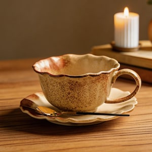 May include: A ceramic teacup and saucer set with a scalloped rim and a speckled, earthy brown finish. A gold spoon rests on the saucer. A lit candle and a small vase with greenery are in the background, creating a cozy atmosphere.