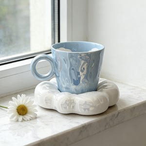 Pode incluir: Uma caneca de cerâmica azul clara com uma superfície texturizada e uma alça redonda está sobre um descanso de copo branco em forma de nuvem. Uma margarida com centro amarelo está por perto. A caneca e o descanso de copo estão em uma janela branca.