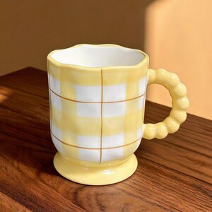 May include: A yellow and white checkered ceramic mug with a unique handle and base. The mug has a wavy rim and a grid pattern. The handle is a series of connected spheres. The mug is sitting on a wooden surface.