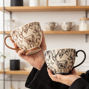 Puede incluir: Dos tazas de café de cerámica con diseños de ciervos y flores. Una taza tiene un asa y una base marrones, la otra tiene un asa y una base negras. Las tazas son beige con ilustraciones marrones y negras.