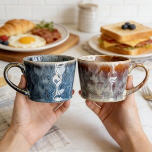 May include: Two ceramic mugs with textured surfaces and curved handles. One mug is blue, the other is brown with a gradient effect. The mugs are held in front of a breakfast setting with food on plates, including a croissant, fried egg, and sandwich.