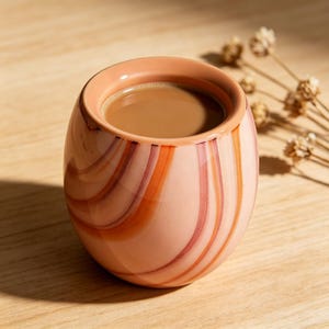 Puede incluir: Una taza de cerámica con un diseño en espiral en tonos melocotón, naranja y burdeos. La taza contiene un líquido marrón claro, probablemente café o té. La taza está sobre una superficie de madera.