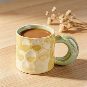 Puede incluir: Una taza de cerámica con un patrón de panal en tonos amarillos y crema. La taza tiene un borde y un asa verdes y está llena de un líquido marrón. La taza está sobre una superficie de madera.