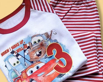 May include: White and red pajama set featuring a graphic of Lightning McQueen and other Cars characters. The shirt has a red neckline and sleeves, with the number 3 and the name "NATE". The pants are red and white striped.