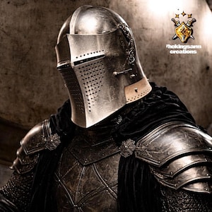 May include: A close-up of a knight's helmet and armor. The silver helmet has a visor with small holes and is part of a full suit of armor. The armor is detailed with intricate patterns and is worn with a black cape. The logo "thekingsarm creations" is in the upper right corner.