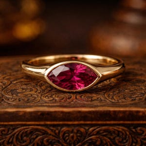 以下が含まれることがあります： 深紅色のマーキス型宝石をあしらったゴールドリング。 リングのバンドは滑らかで磨かれており、宝石はベゼルセッティングでセットされています。 リングは木製の表面に展示されており、そのエレガントなデザインを際立たせています。