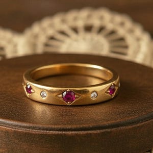 Peut inclure: Une bague en or ornée de pierres précieuses rouges et claires alternées. La bague comporte trois pierres rouges et deux pierres claires. Elle est présentée sur un support circulaire marron, avec une nappe en dentelle en arrière-plan.