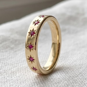May include: A gold band ring featuring a series of small, round, deep pink rubies set into the metal. Each ruby is surrounded by an engraved starburst pattern, creating a repeating celestial motif along the outer surface of the polished gold ring.
